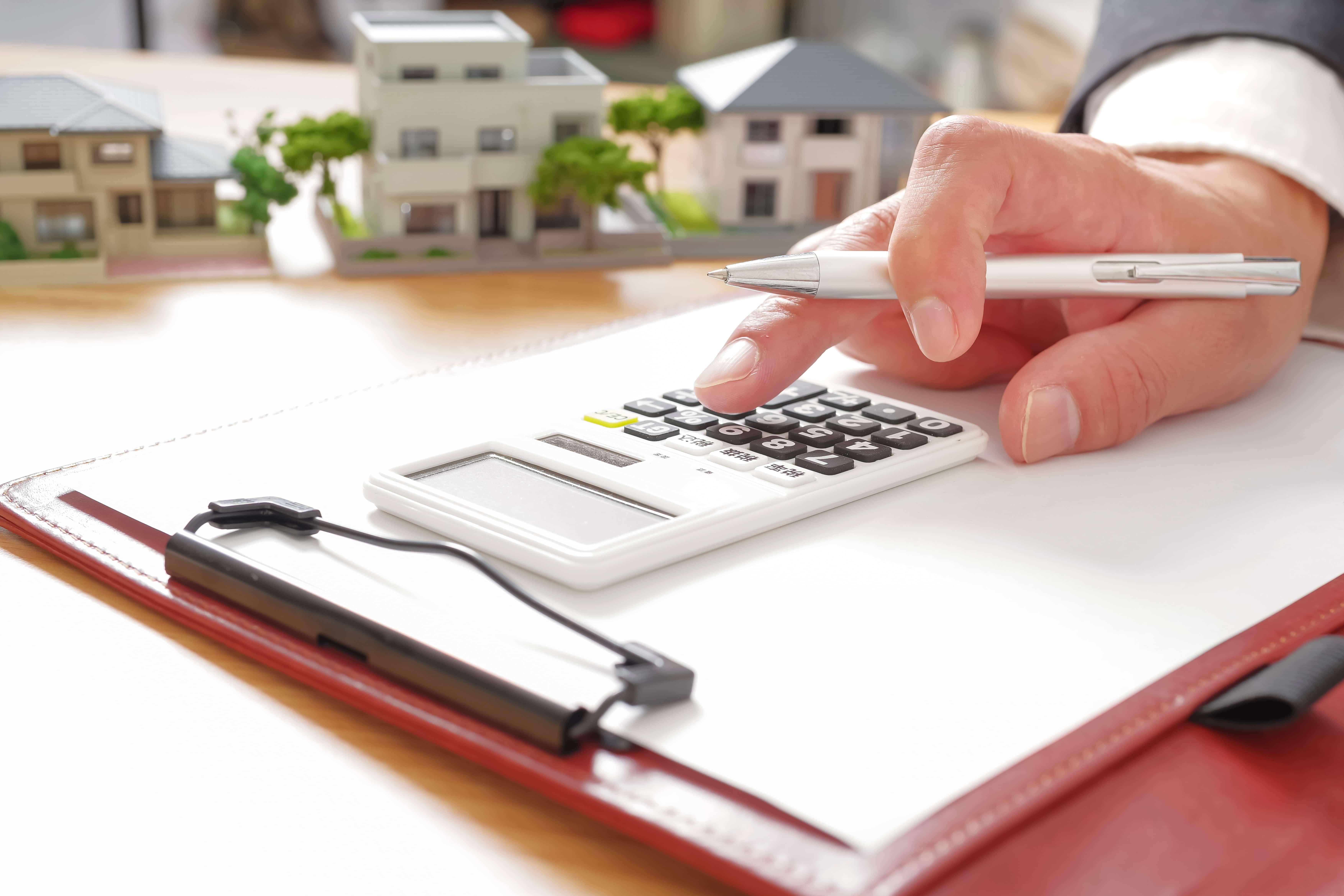 電卓を操作する手と赤いバインダー、背景に並ぶ住宅ミニチュア模型