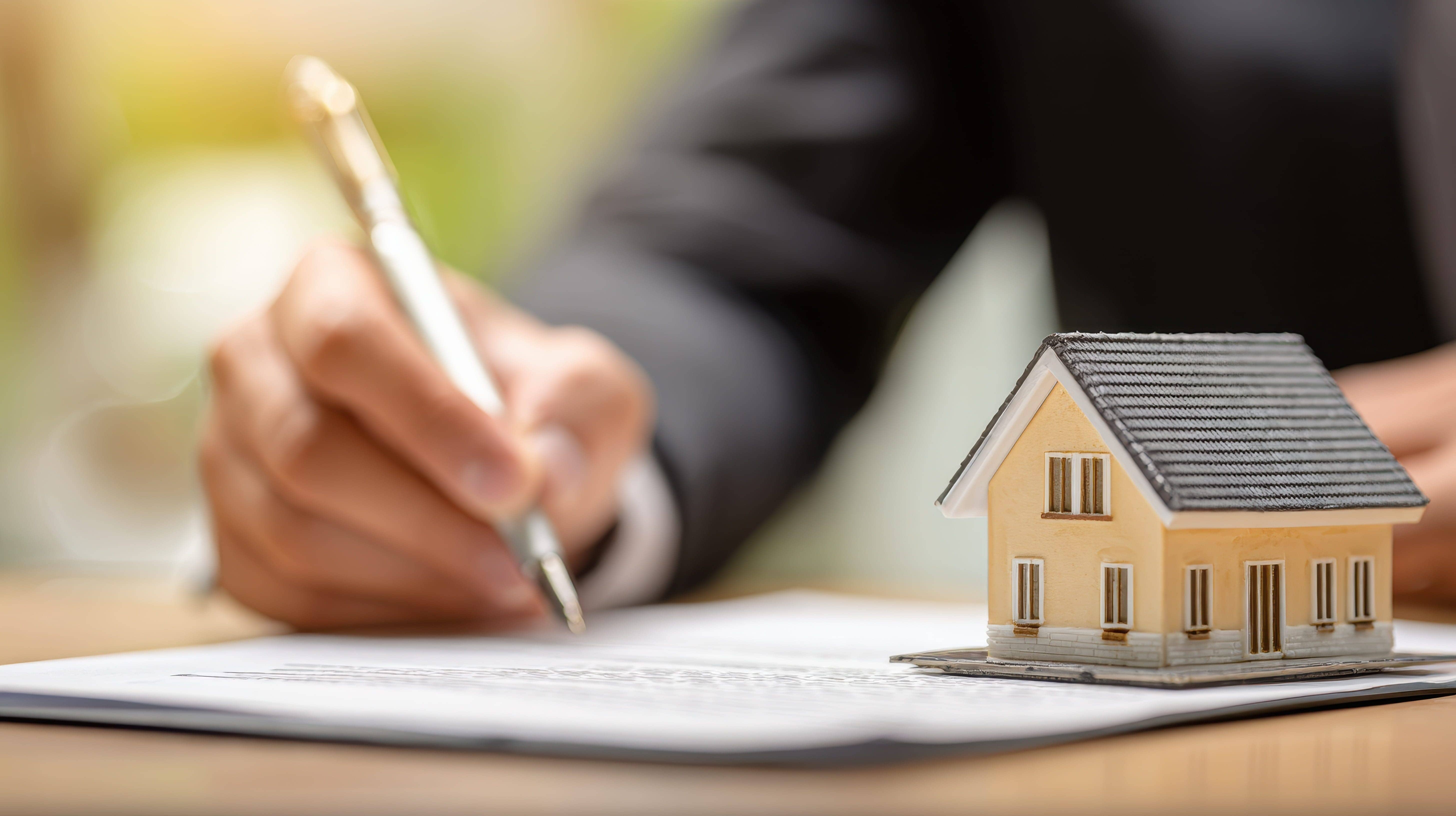 スーツ姿の人が書類にサインする様子と、手前に置かれたベージュの住宅ミニチュア模型